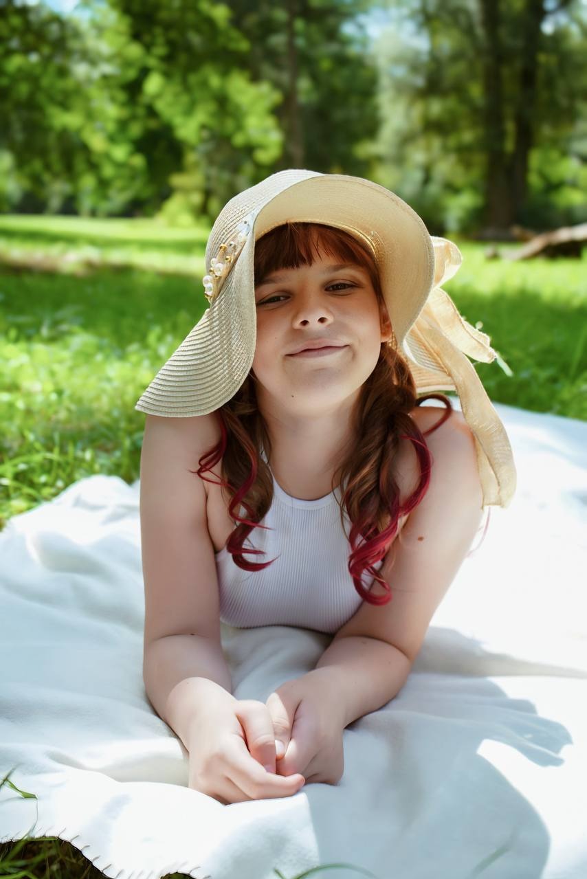 Portret młodej kobiety w kapeluszu, leżącej na kocu w parku, z delikatnym uśmiechem i naturalnym oświetleniem, podkreślającym jej młodzieńczy wygląd.