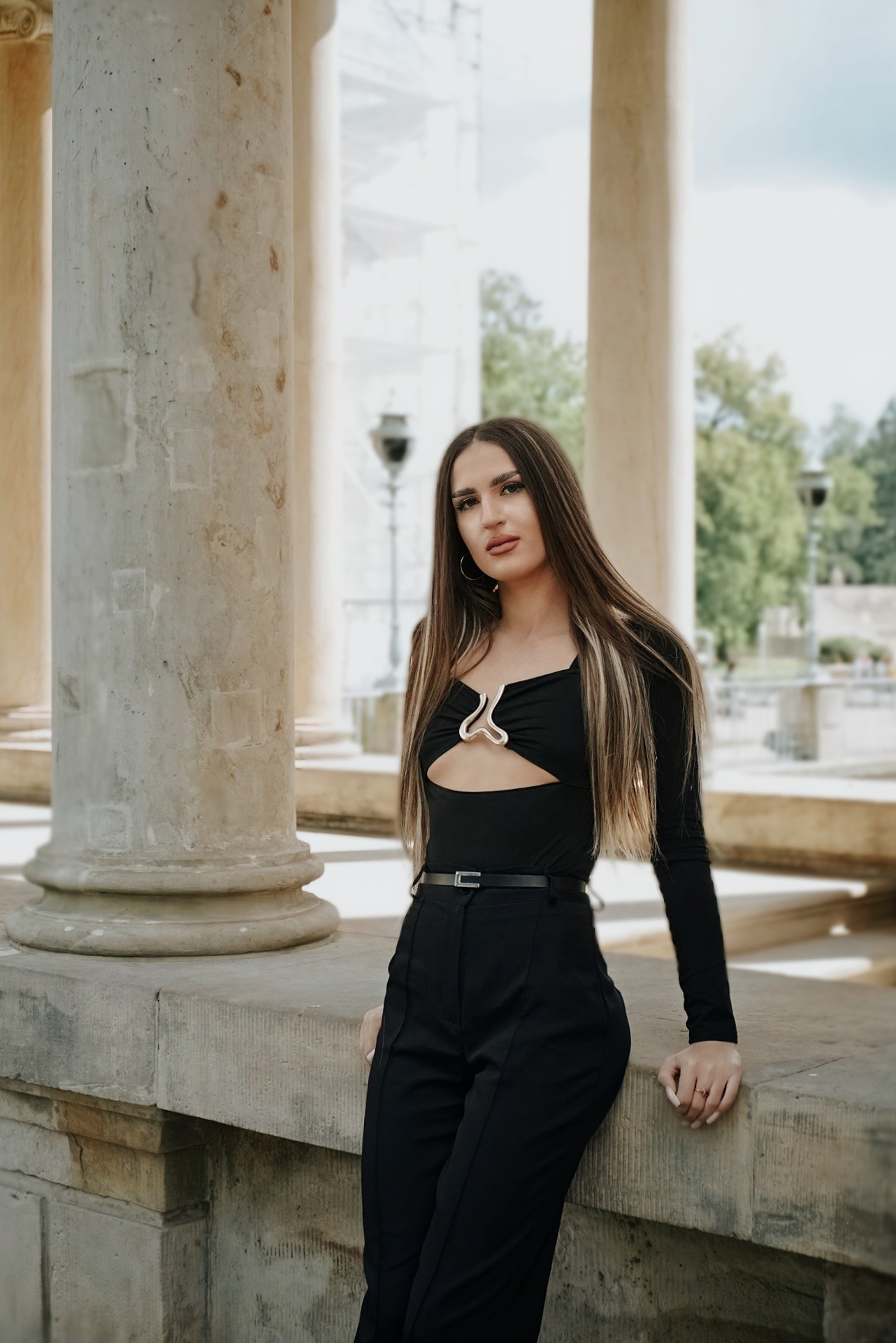 Portret kobiety w czarnym stroju, opierającej się o kamienny murek z kolumnami w tle, w plenerze. Ujęcie z delikatnym rozmyciem tła.