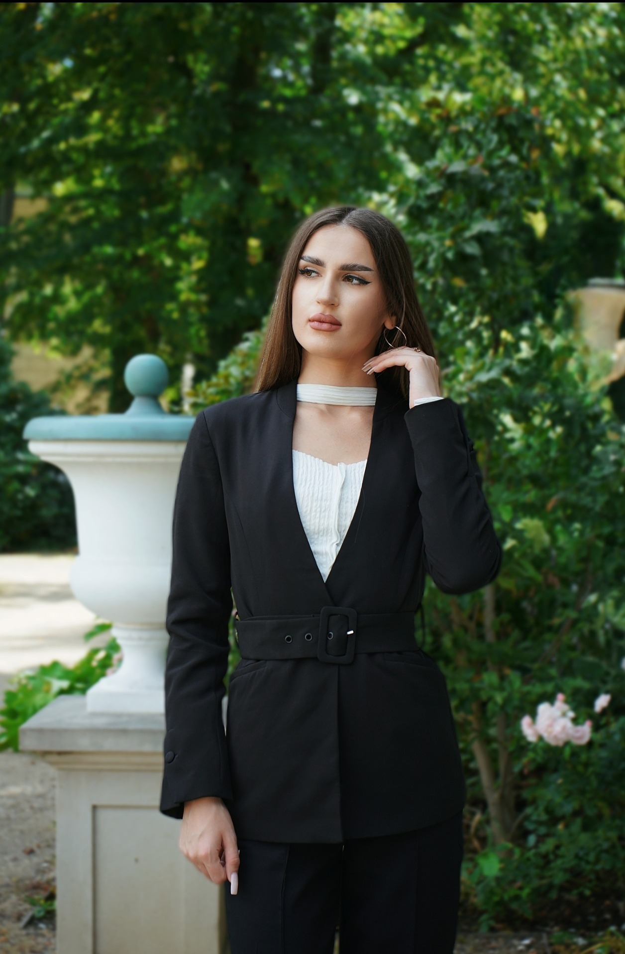 Kobieta w czarnym garniturze pozuje na tle zieleni, z ręką uniesioną do szyi. Widoczna architektura parkowa i kwiaty w tle. Subtelna biżuteria i makijaż.