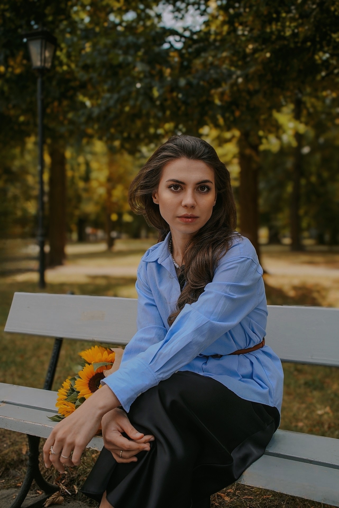 Portret kobiety w błękitnej koszuli siedzącej na ławce w parku z bukietem słoneczników. Jesienna sceneria, miękkie światło, naturalny wyraz twarzy.