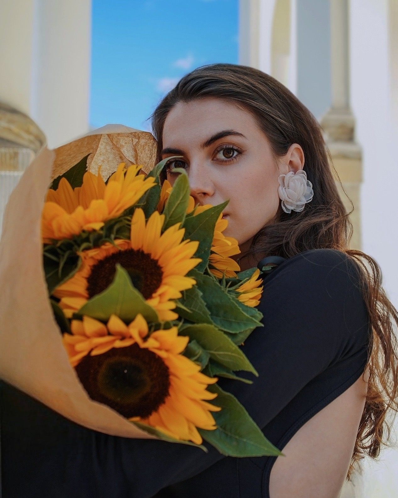 Portret kobiety z bukietem słoneczników, z kolczykiem w kształcie kwiatu na tle kolumn i błękitnego nieba, z delikatnym makijażem i długimi włosami.