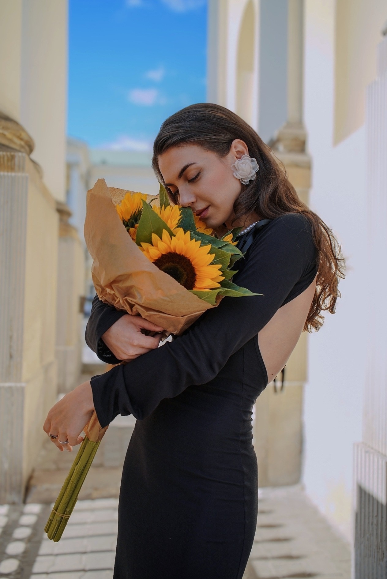 Elegancka brunetka w czarnej sukni z odkrytymi plecami, tuląca bukiet słoneczników na tle kolumnady w słoneczny dzień. Delikatne kolczyki w kształcie kwiatów.