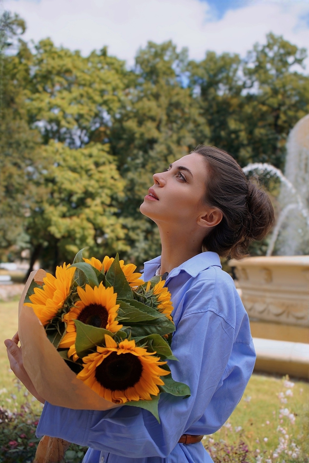 Portret kobiety z bukietem słoneczników w plenerze, spoglądającej w górę. W tle fontanna i zieleń parku, ciepłe światło słoneczne. Ujęcie z dołu.