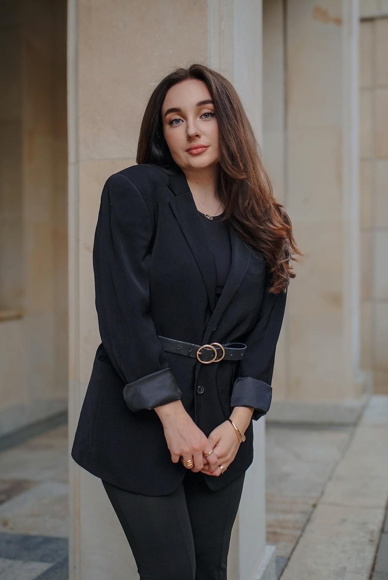 Portret kobiety w czarnym garniturze z paskiem, stojącej na tle kolumn. Uwagę przykuwają złote pierścionki i bransoletka. Stonowana kolorystyka i miękkie światło.