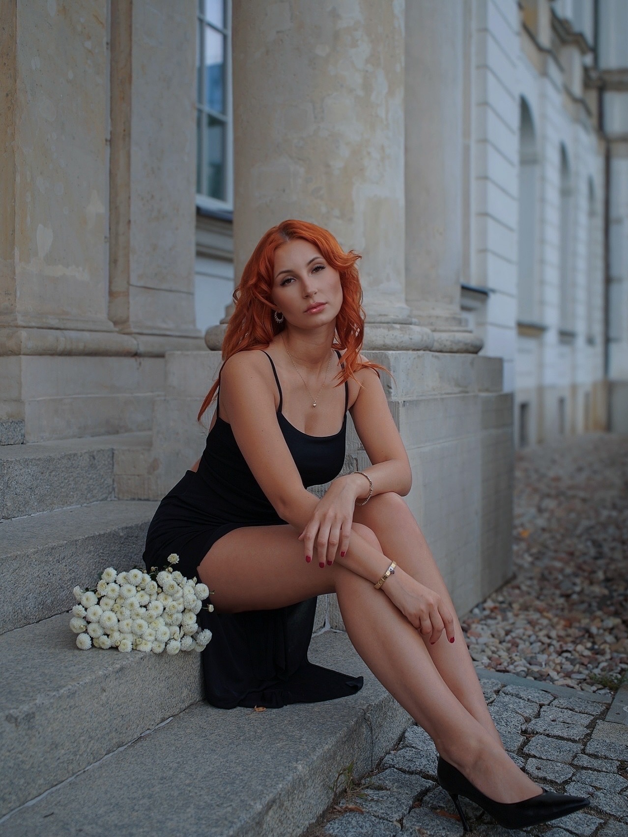 Kobieta z rudymi włosami w czarnej sukience siedzi na schodach kolumnady z bukietem białych kwiatów. Portret w stonowanej kolorystyce.
