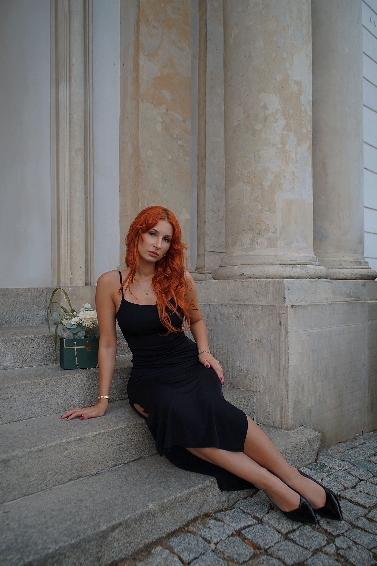 Portret kobiety z rudymi włosami w czarnej sukience, siedzącej na schodach z kwiatami w ozdobnym pudełku, na tle kolumn. Sesja w plenerze.