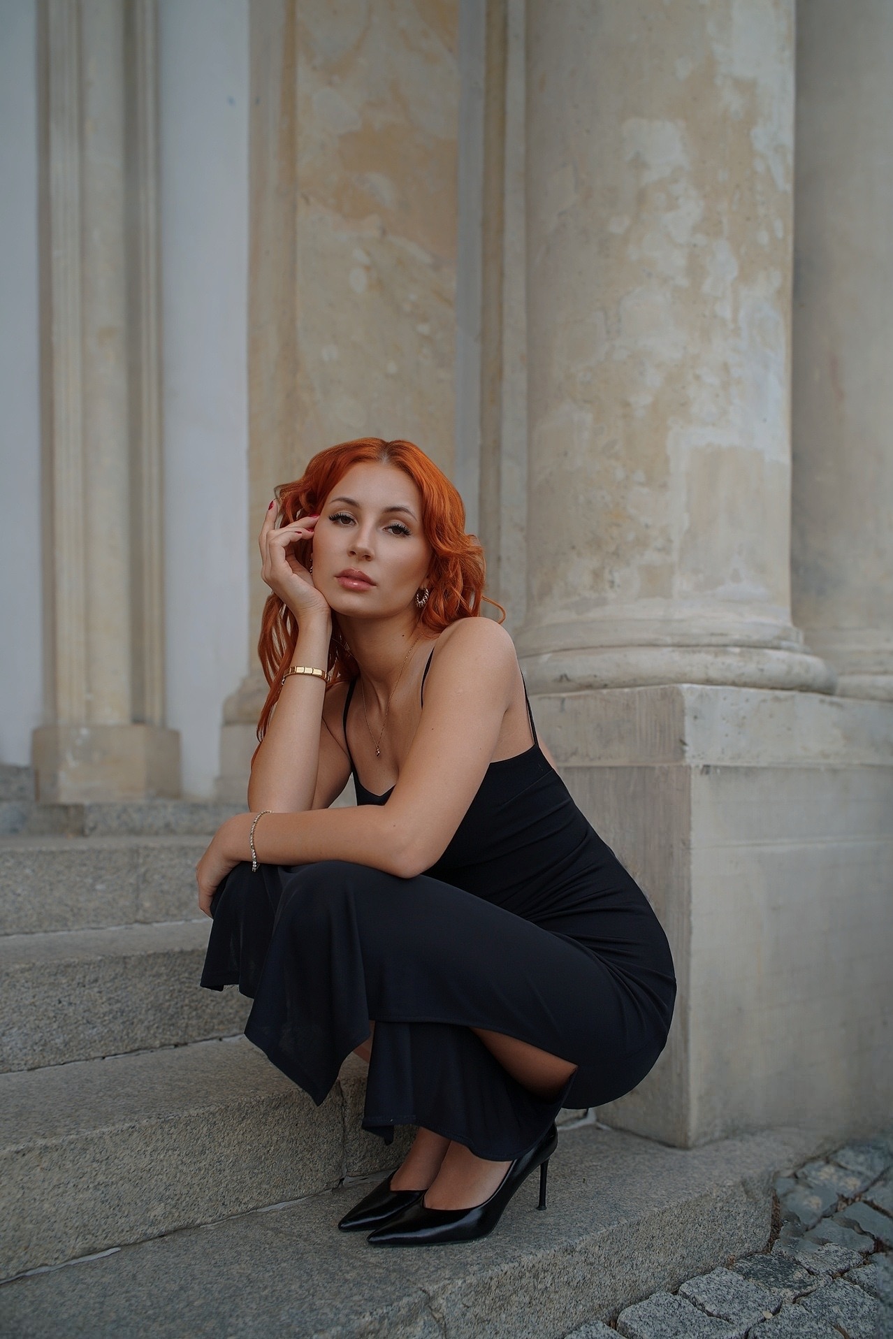 Portret kobiety z rudymi włosami w czarnej sukience, siedzącej na schodach kolumnady. Elegancki strój i subtelna biżuteria podkreślają profesjonalny charakter.