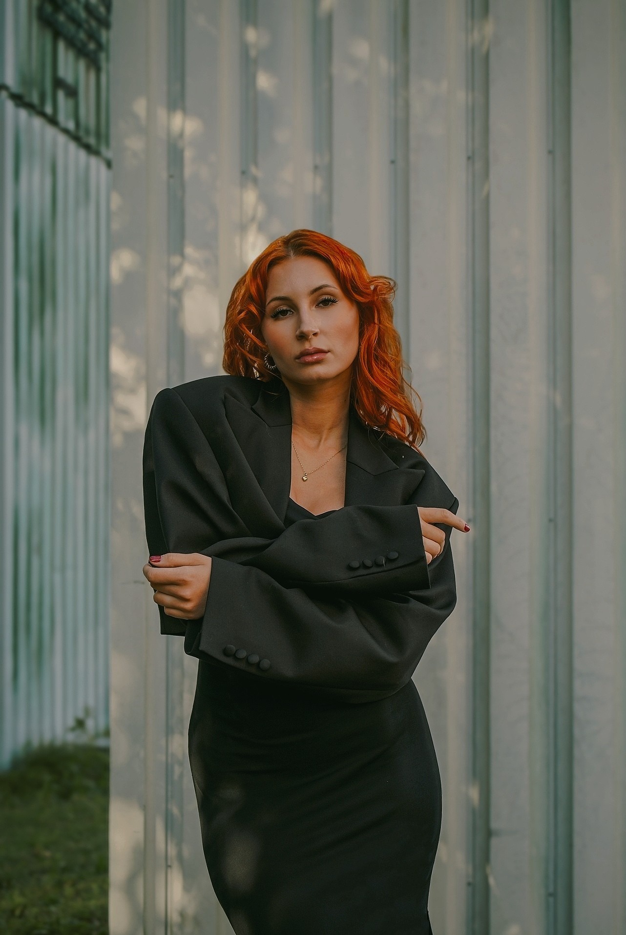 Kobieta o rudych włosach w czarnym, oversize'owym garniturze na tle blachy falistej w cieniu. Minimalistyczna biżuteria i profesjonalny makijaż. Całość w stonowanej kolorystyce.