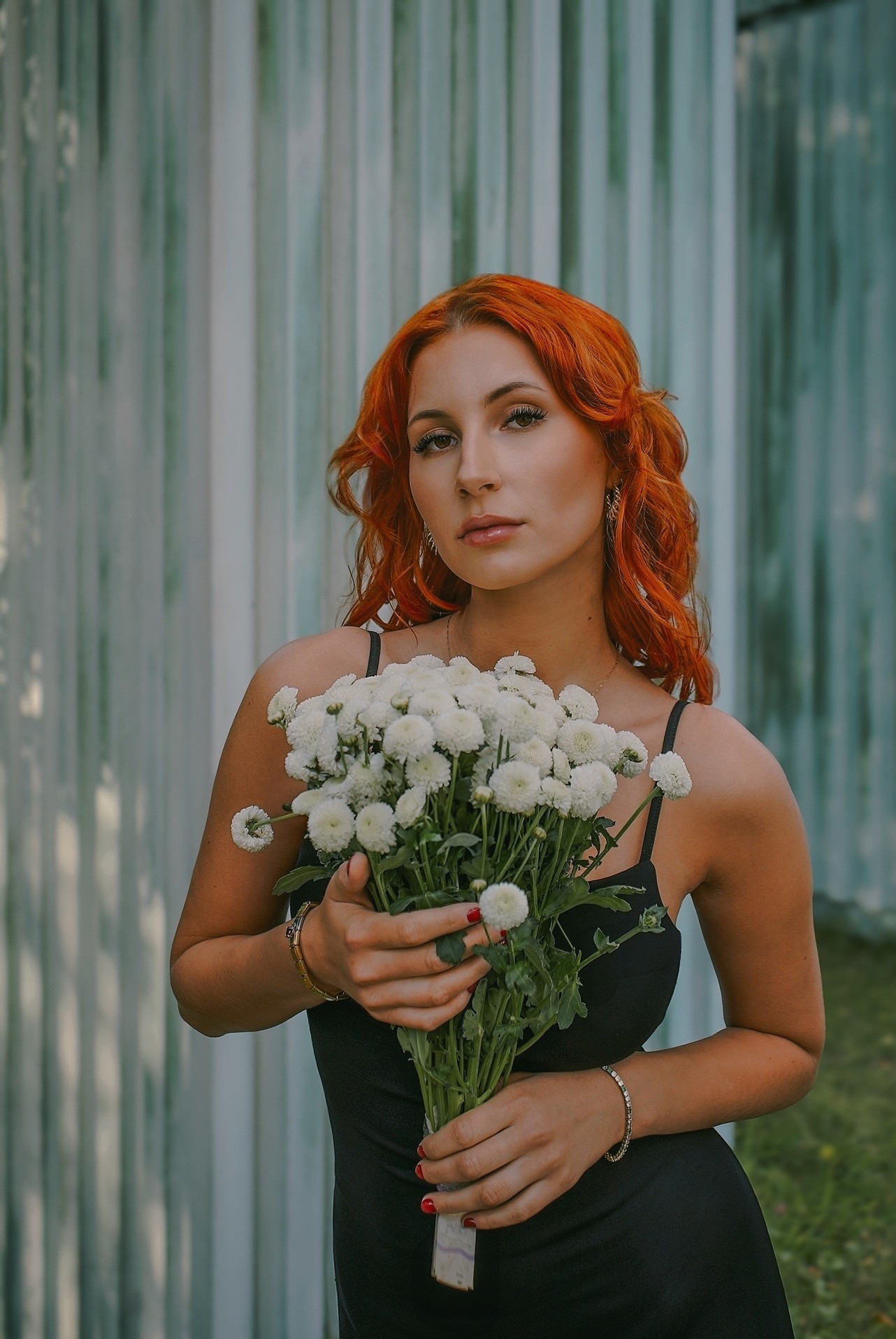 Portret kobiety o rudych włosach, trzymającej bukiet białych chryzantem na tle blaszanej ściany. Ubrana w czarną sukienkę na ramiączkach, z delikatną biżuterią.