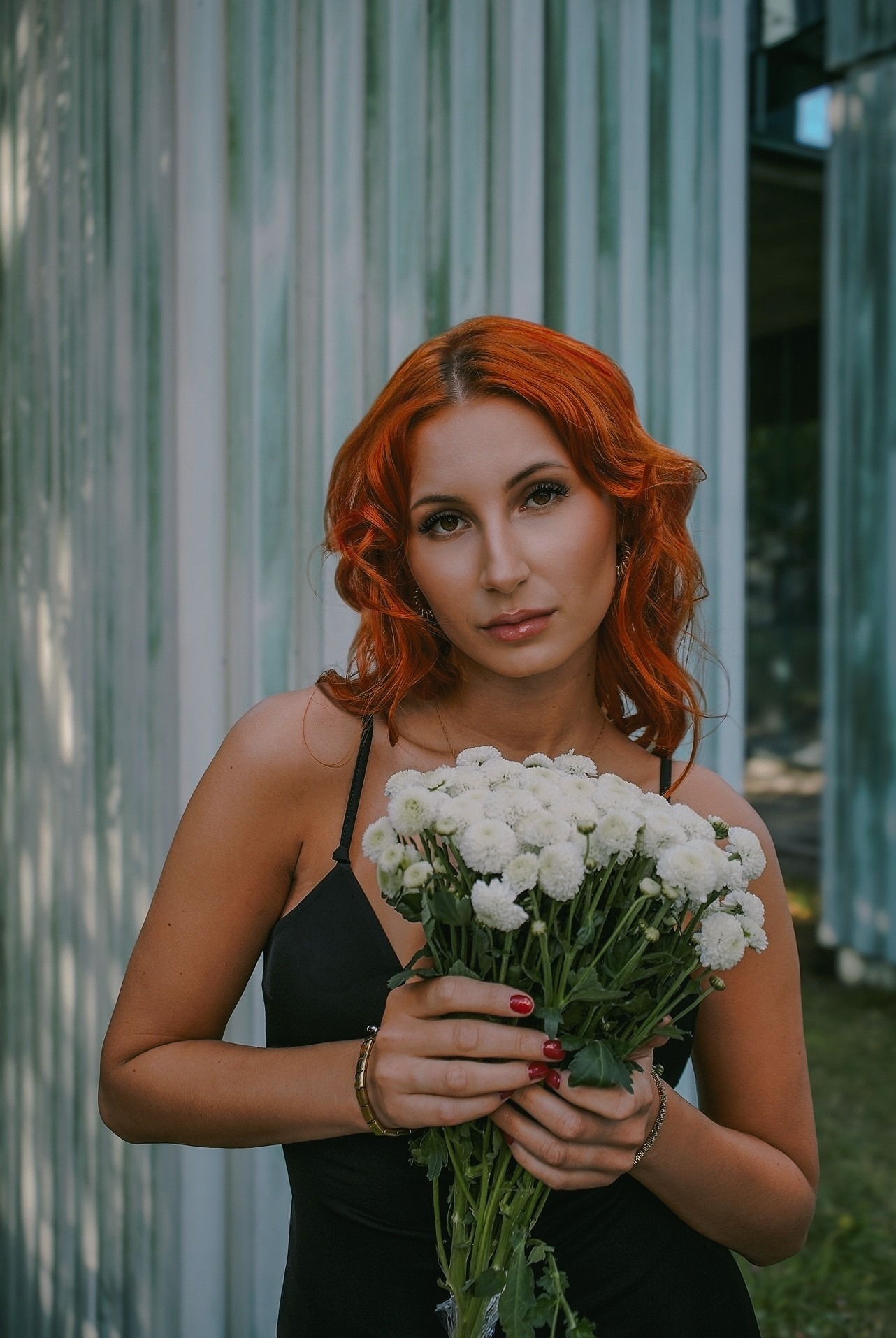 Portret kobiety z rudymi włosami, trzymającej bukiet białych kwiatów na tle metalowej konstrukcji. Ubrana w czarną sukienkę na ramiączkach, z delikatnym makijażem i biżuterią.