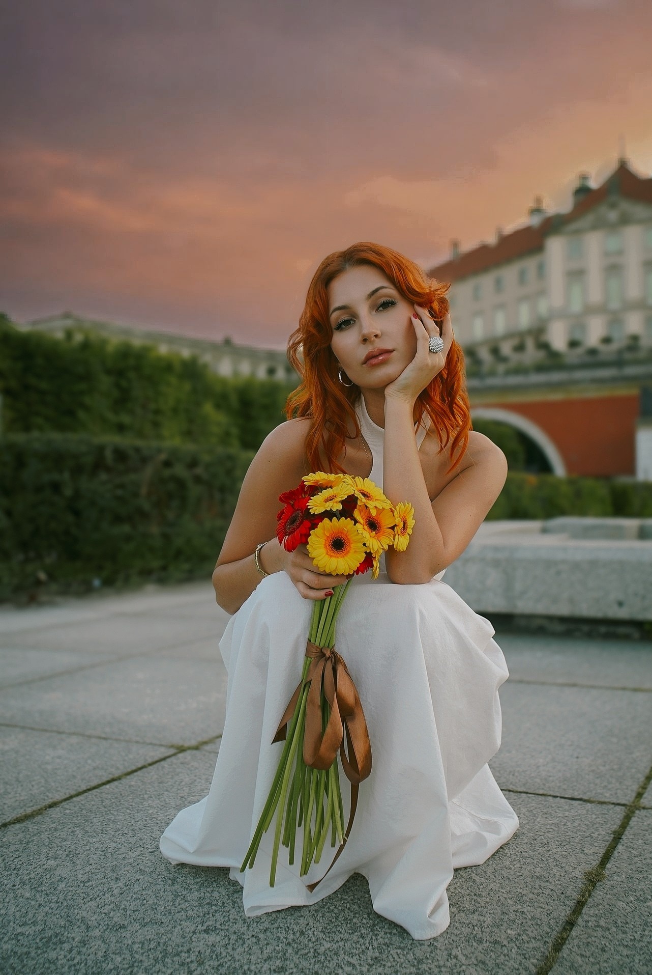 Portret kobiety z rudymi włosami w białej sukience, trzymającej bukiet gerber, na tle architektury i zachodzącego słońca. Uwagę przykuwa duży pierścionek.
