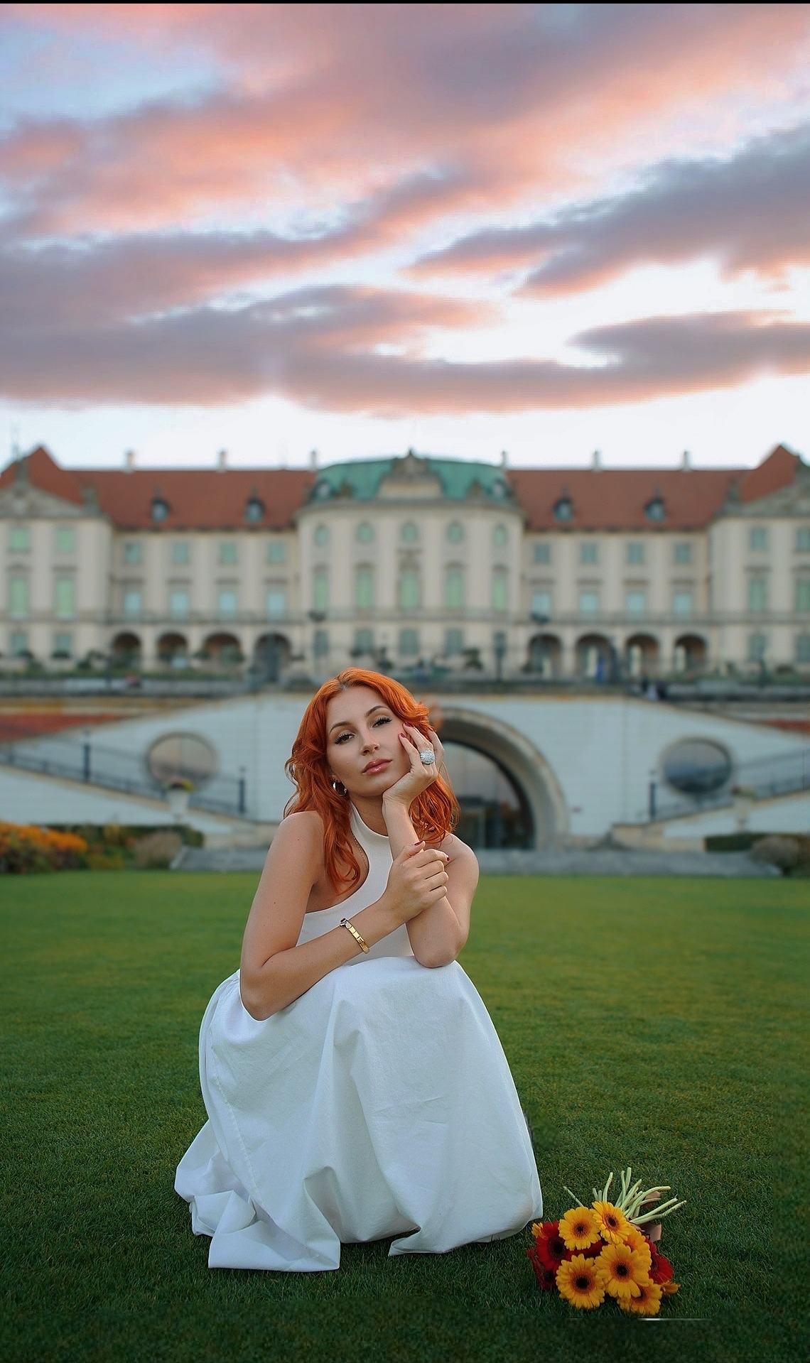 Kobieta w białej sukni klęczy na trawie z bukietem, na tle zamku i zachodzącego słońca. Ujęcie z niskiego kąta podkreśla architekturę i niebo.