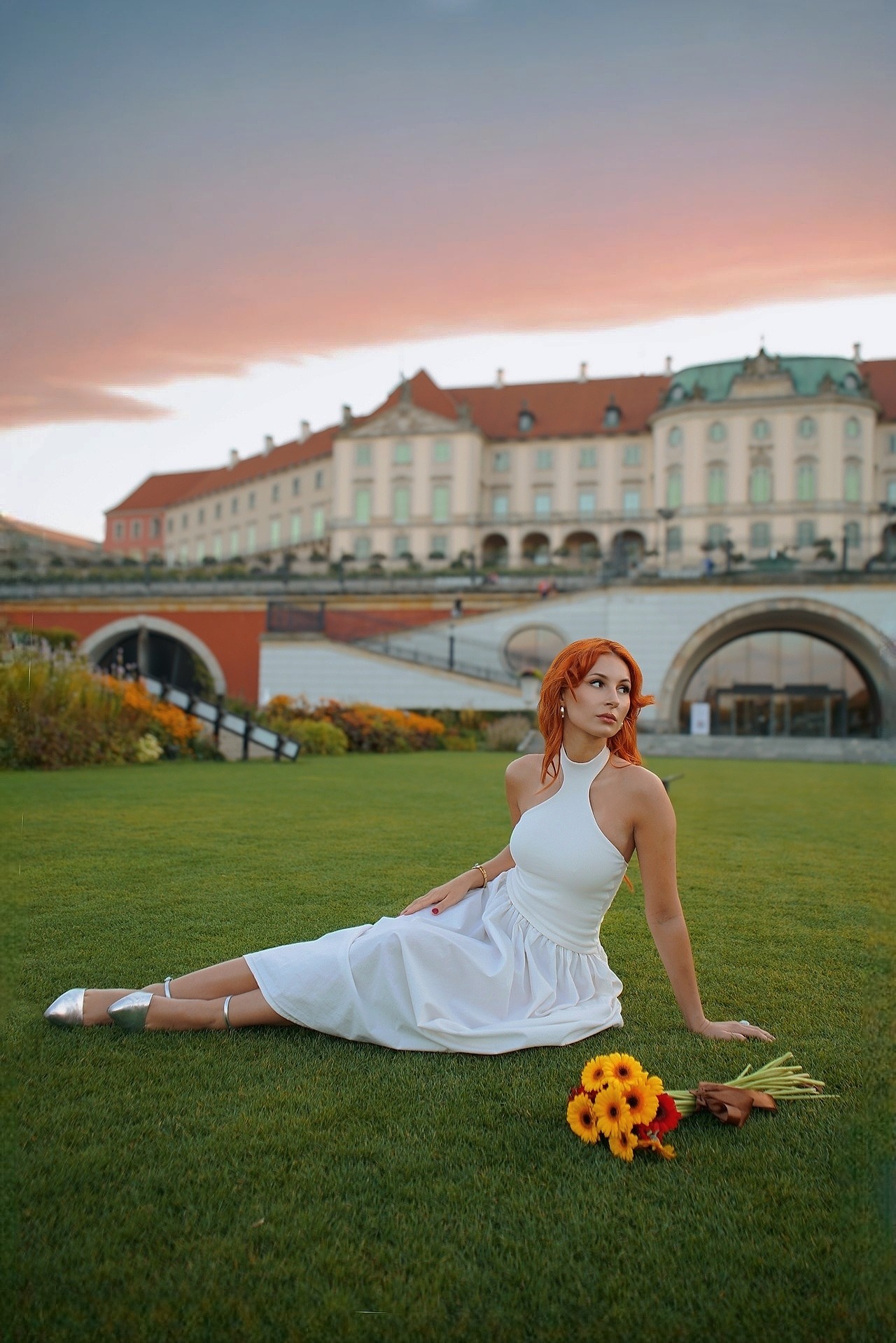 Kobieta w białej sukience leży na trawie z bukietem słoneczników, w tle zamek i zachodzące słońce. Ujęcie z dołu, podkreślające sylwetkę i architekturę.