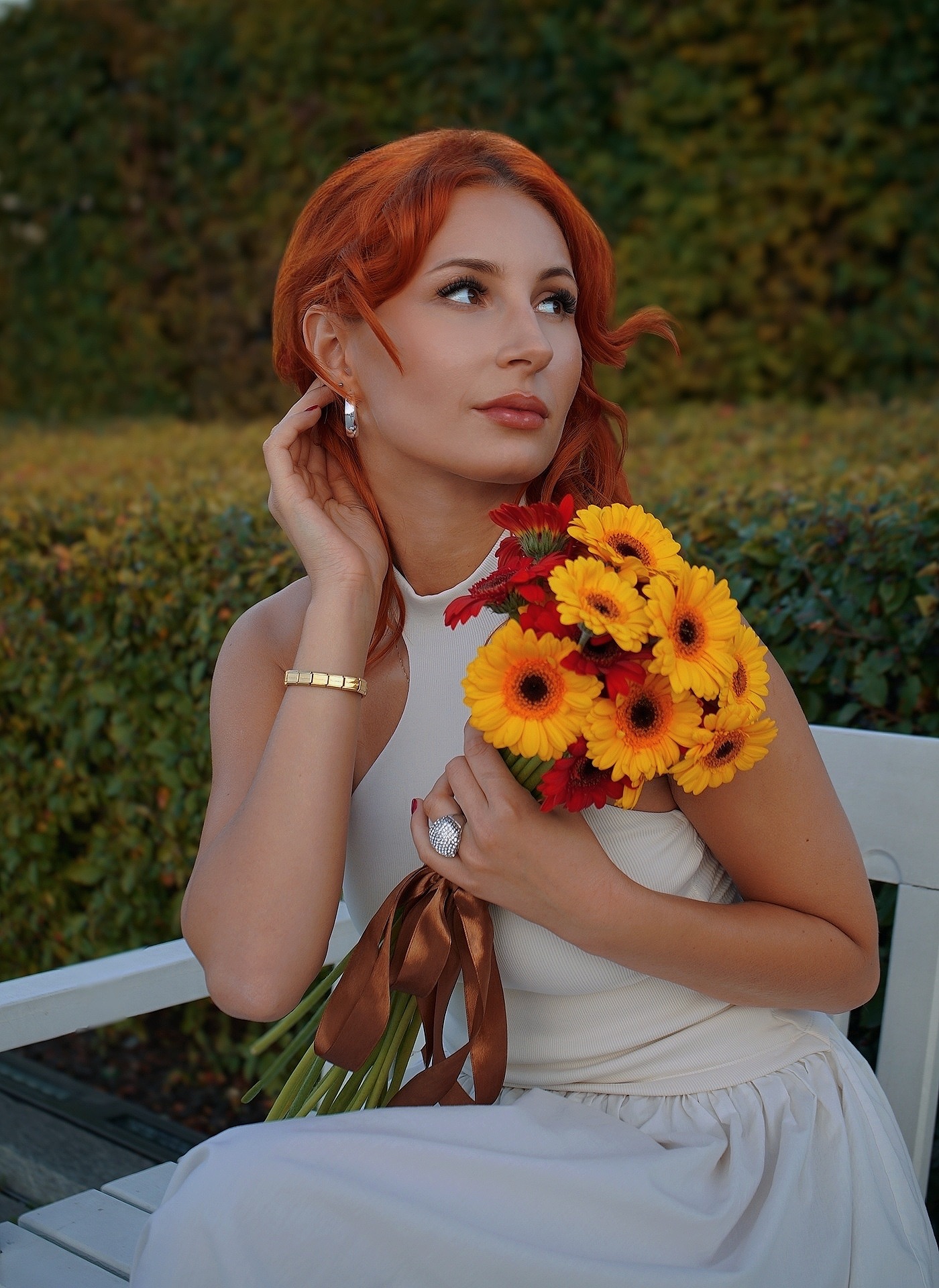 Portret kobiety z rudymi włosami, trzymającej bukiet gerber w odcieniach żółci i czerwieni, siedzącej na białej ławce na tle zielonych krzewów.