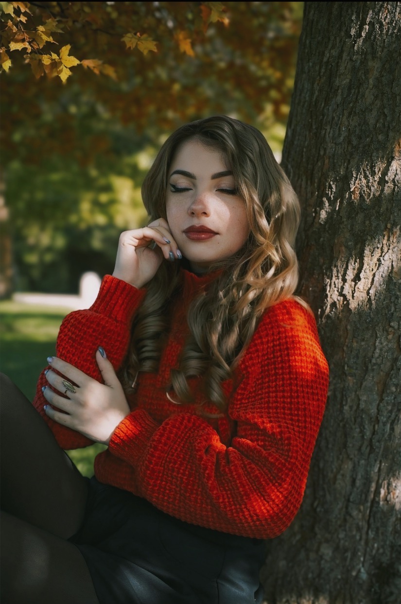 Portret kobiety w czerwonym swetrze, opierającej się o drzewo w jesiennym parku, z zamkniętymi oczami i delikatnym uśmiechem, w naturalnym świetle słonecznym.