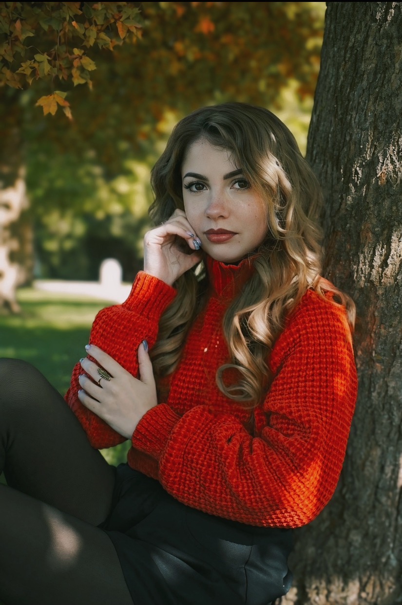 Portret młodej kobiety w czerwonym swetrze, opierającej się o drzewo w jesiennej scenerii, z delikatnym uśmiechem i subtelnym makijażem.