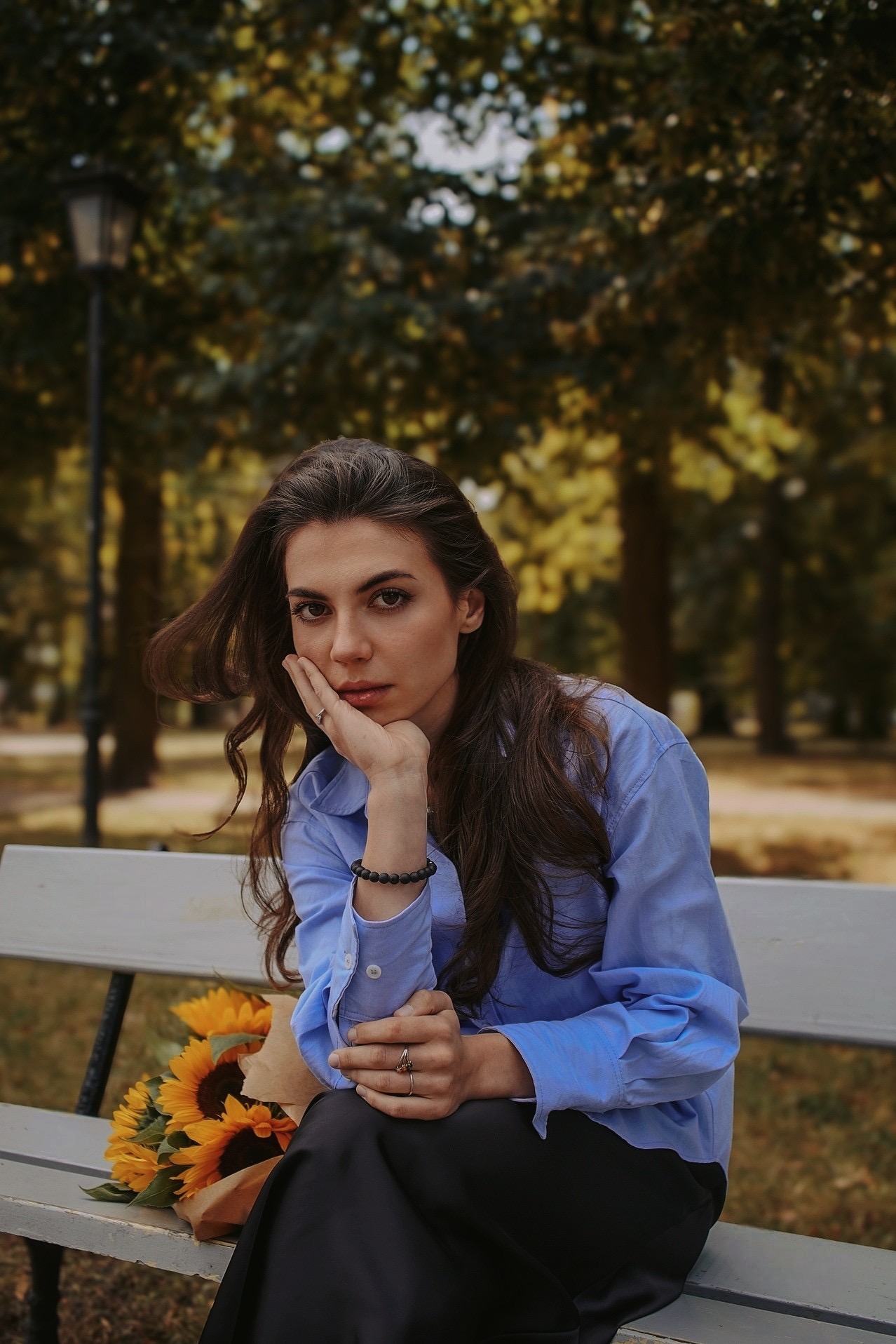 Portret kobiety w błękitnej koszuli, siedzącej na ławce z bukietem słoneczników, na tle parku w jesiennej scenerii. Delikatne światło podkreśla urodę modelki.