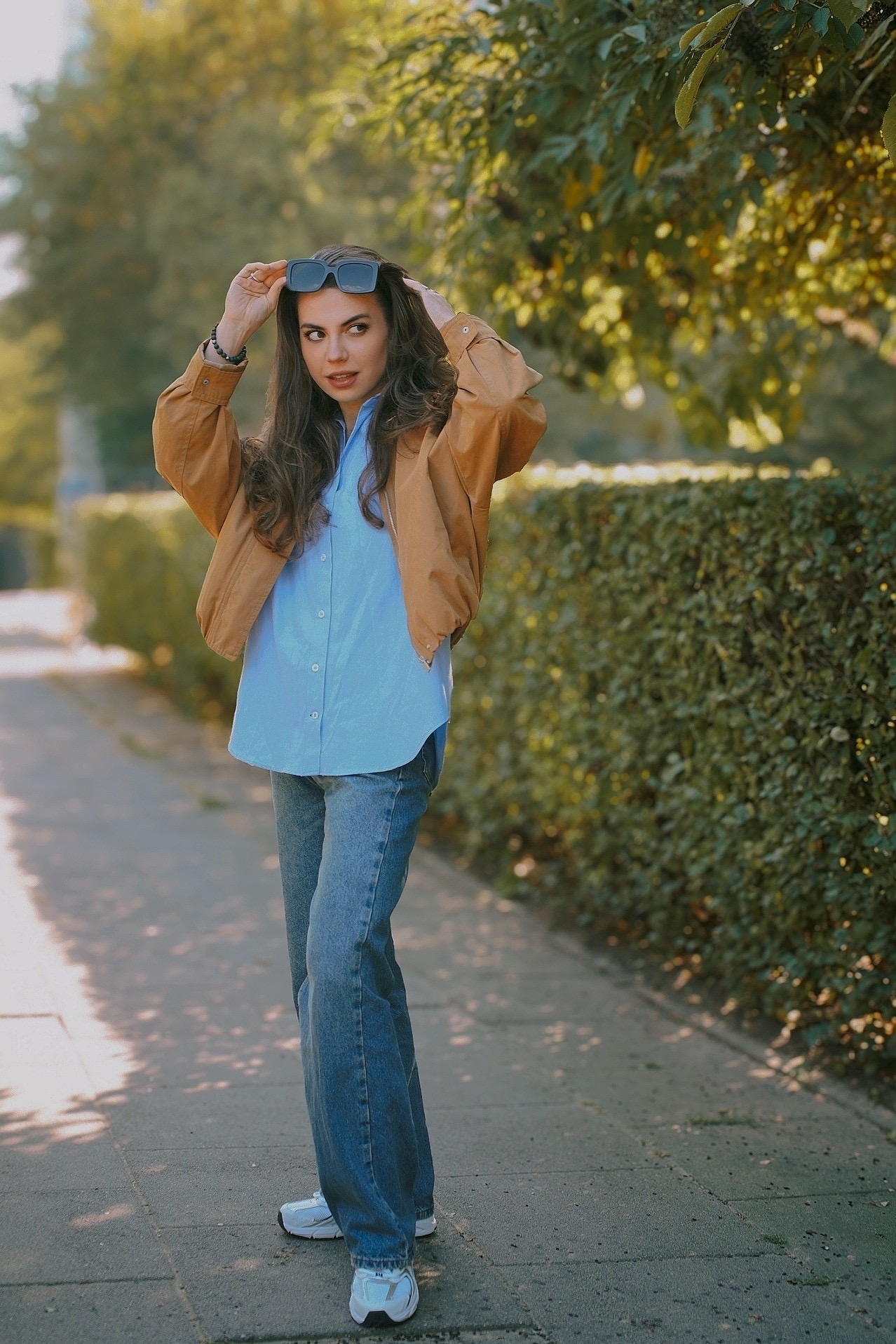 Kobieta w brązowej kurtce i jeansach pozuje na chodniku, trzymając okulary na głowie. Za nią żywopłot i drzewa w słoneczny dzień.