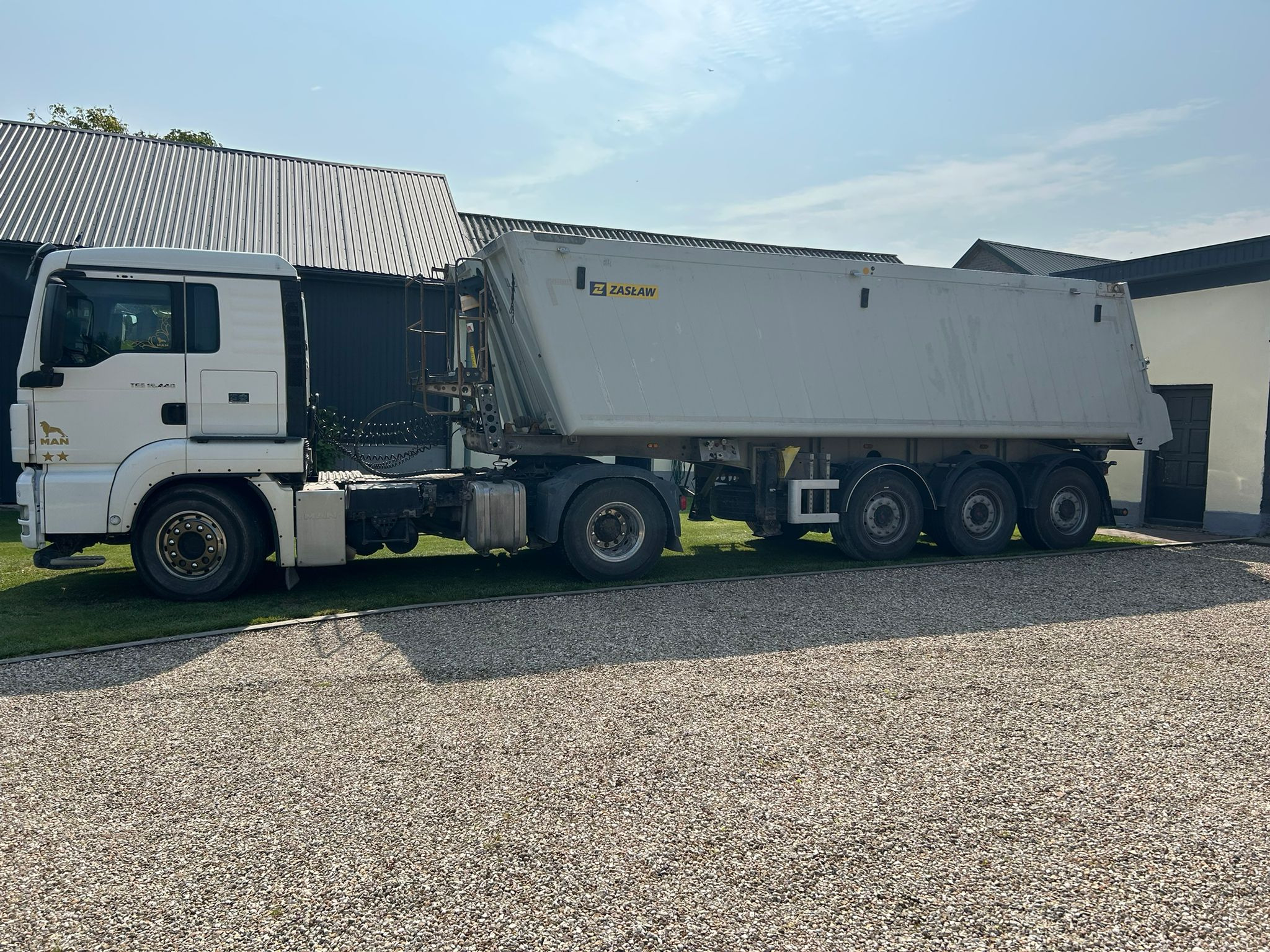 Ciężarówka MAN z naczepą ZASŁAW na żwirowym podłożu, widok z boku, w tle budynki o metalowych dachach. Dzienny kadr prezentuje transport ciężarowy.
