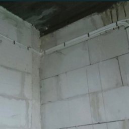 Usługi Elektryczne - Białe kable elektryczne prowadzone wzdłuż surowych ścian z betonu komórkowego, zabezpieczone czarnymi uchwytami. Widok na narożnik pomieszczenia w trakcie prac instalacyjnych.