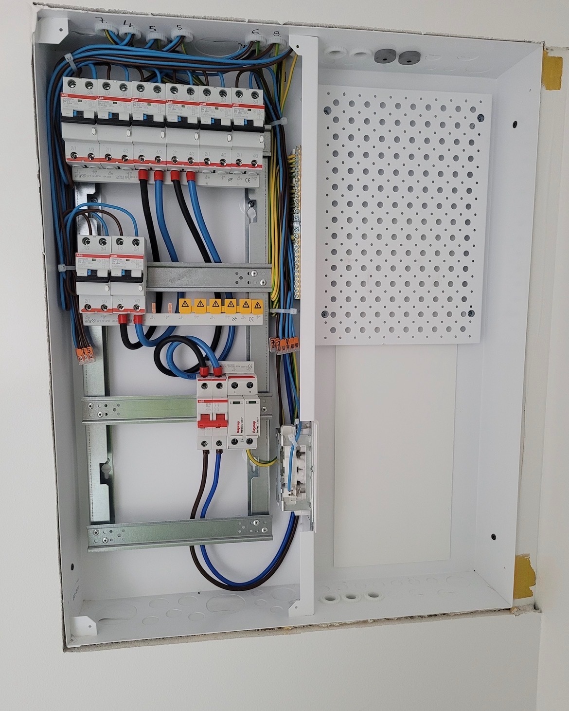 Rozdzielnica elektryczna w białej obudowie, widoczne zabezpieczenia, przewody w kolorach niebieskim, czarnym i żółto-zielonym. Elementy montażowe i perforowana płyta w tle.