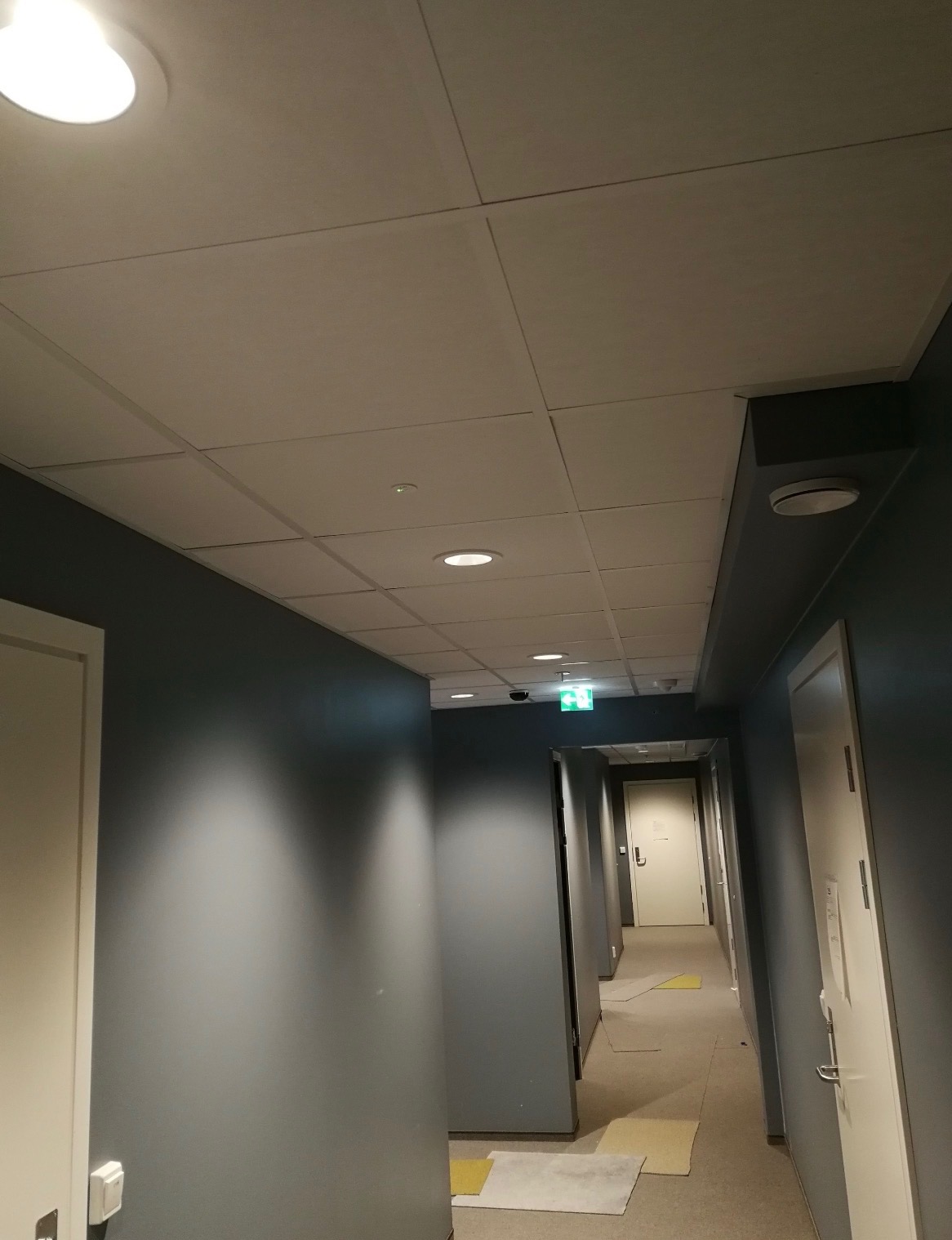 Długi korytarz z szarą ścianą i białymi drzwiami, oświetlony okrągłymi lampami sufitowymi i zielonym znakiem ewakuacyjnym. Widok z dołu na sufit kasetonowy.