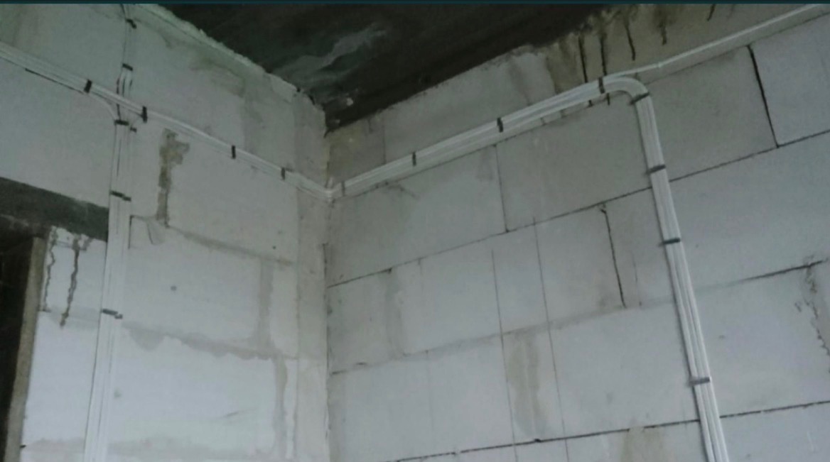 Białe kable elektryczne prowadzone wzdłuż surowych ścian z betonu komórkowego, zabezpieczone czarnymi uchwytami. Widok na narożnik pomieszczenia w trakcie prac instalacyjnych.