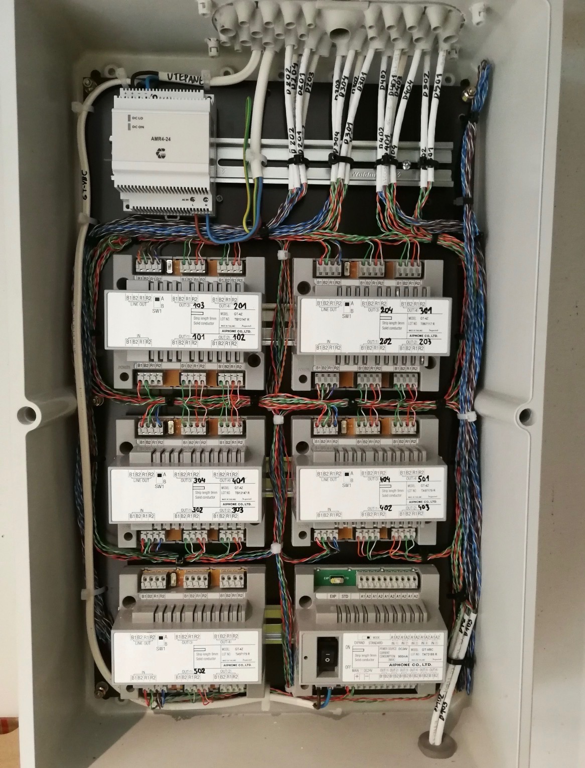 Rozdzielnica domofonowa marki Aiphone z licznymi przewodami i modułami, zamontowana w skrzynce. Widoczne oznaczenia i numery na modułach. Estetyczne ułożenie kabli.