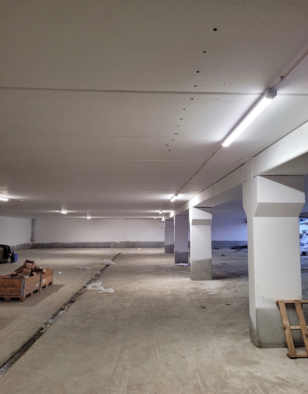 Wnętrze hali w trakcie prac wykończeniowych z widocznymi lampami sufitowymi, betonowymi słupami i elementami instalacji pod sufitem. Widoczne ślady po instalacjach.