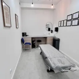 Jasne wnętrze gabinetu z leżanką przykrytą jednorazowym podkładem, wiszącymi lampami i ścianą z certyfikatami. Minimalistyczny wystrój, szare płytki podłogowe.