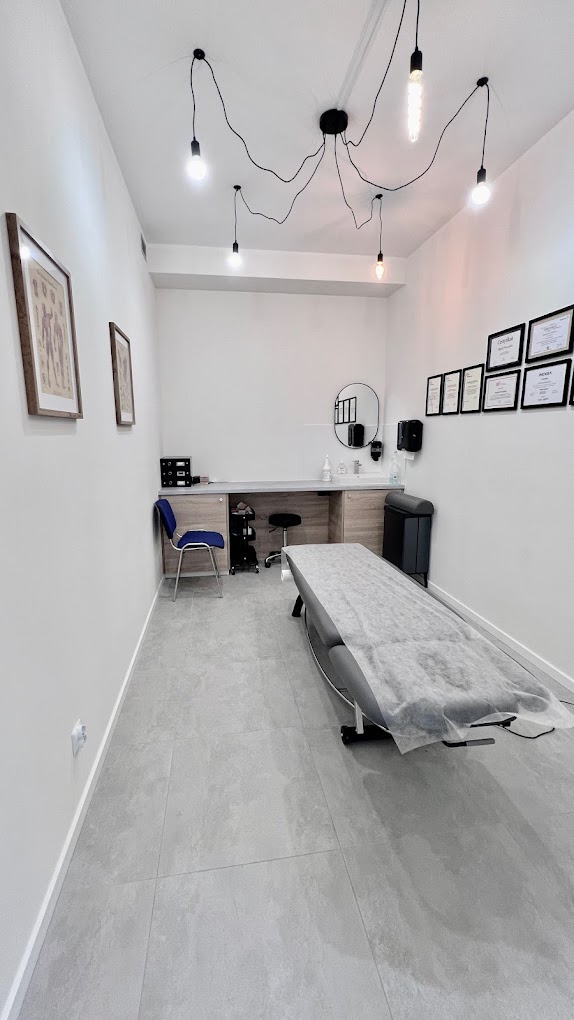 Jasne wnętrze gabinetu z leżanką przykrytą jednorazowym podkładem, wiszącymi lampami i ścianą z certyfikatami. Minimalistyczny wystrój, szare płytki podłogowe.