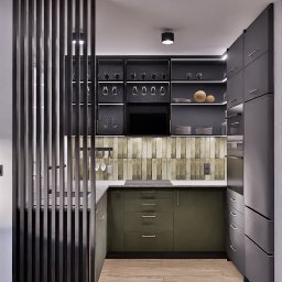 To Oni Projektują - Nowoczesna kuchnia z drewnianą podłogą i zielonymi szafkami, oddzielona od reszty pomieszczenia czarną, ażurową ścianką działową. Minimalistyczne oświetlenie.