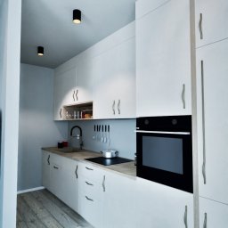 To Oni Projektują - Nowoczesna, minimalistyczna kuchnia z białymi szafkami, drewnianym blatem i podłogą. Czarny piekarnik w zabudowie. Oświetlenie punktowe sufitowe.