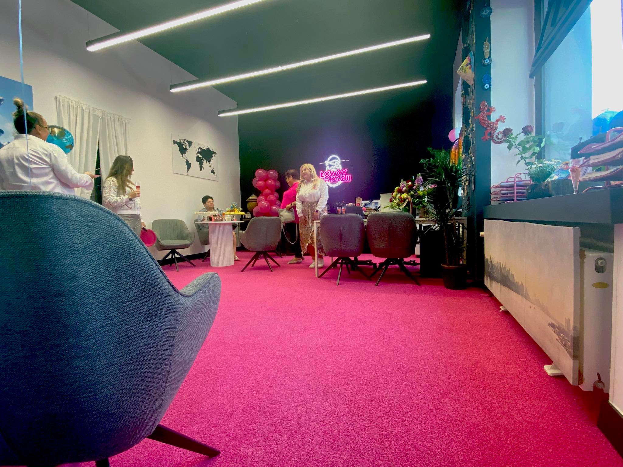 Jasne biuro podróży z różową wykładziną, szarymi krzesłami i neonem. Widoczne osoby, dekoracje i mapa świata na ścianie. Nietypowe połączenie kolorów.