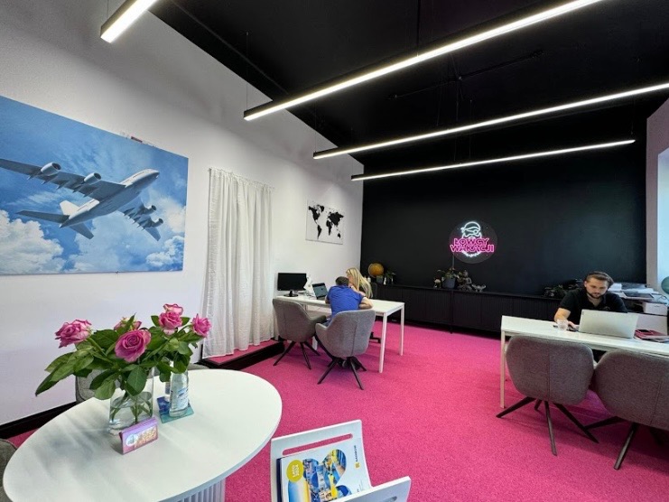 Nowoczesne biuro podróży z różową wykładziną, czarnym sufitem i ścianą. Na ścianie grafika z samolotem i neon 'Łowcy Wakacji'. Osoby pracują przy biurkach.