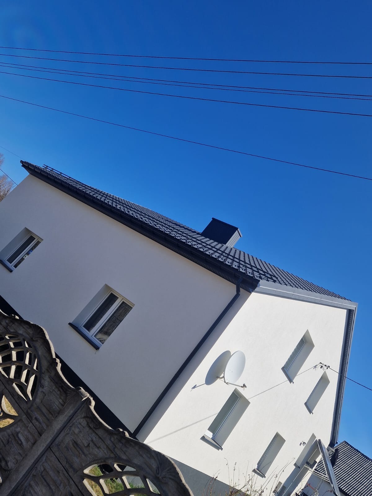 Biały dom z ciemną dachówką widziany z dołu, z fragmentem ozdobnego ogrodzenia. Błękitne niebo w tle, widoczne przewody elektryczne.