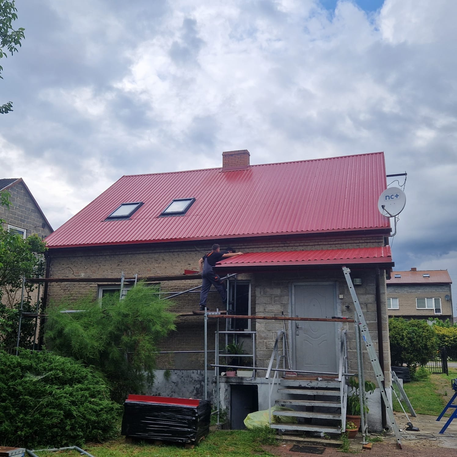 Czerwony dach z blachy falistej na parterowym domu z cegły, widoczne okna dachowe i pracownik na rusztowaniu, montujący obróbki blacharskie.