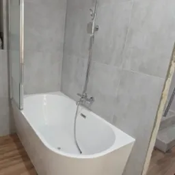 Nowoczesna, biała wanna z chromowaną armaturą i szklanym parawanem na tle szarych płytek. Podłoga wyłożona panelami imitującymi drewno. Widoczny fragment niezakończonej ściany.