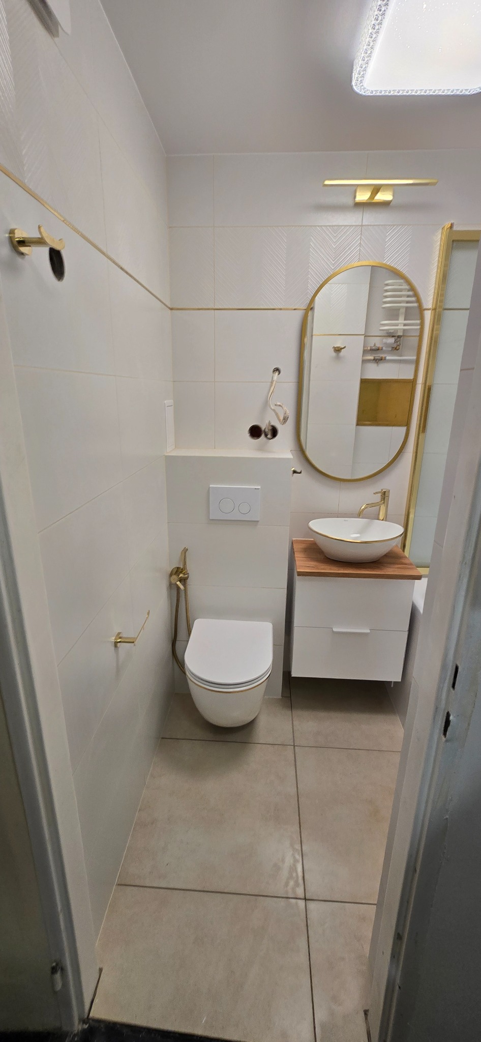Biała łazienka z elementami złota: owalne lustro, umywalka nablatowa na drewnianym blacie, wisząca toaleta. Widoczne białe płytki z delikatnym wzorem i złotymi akcentami.