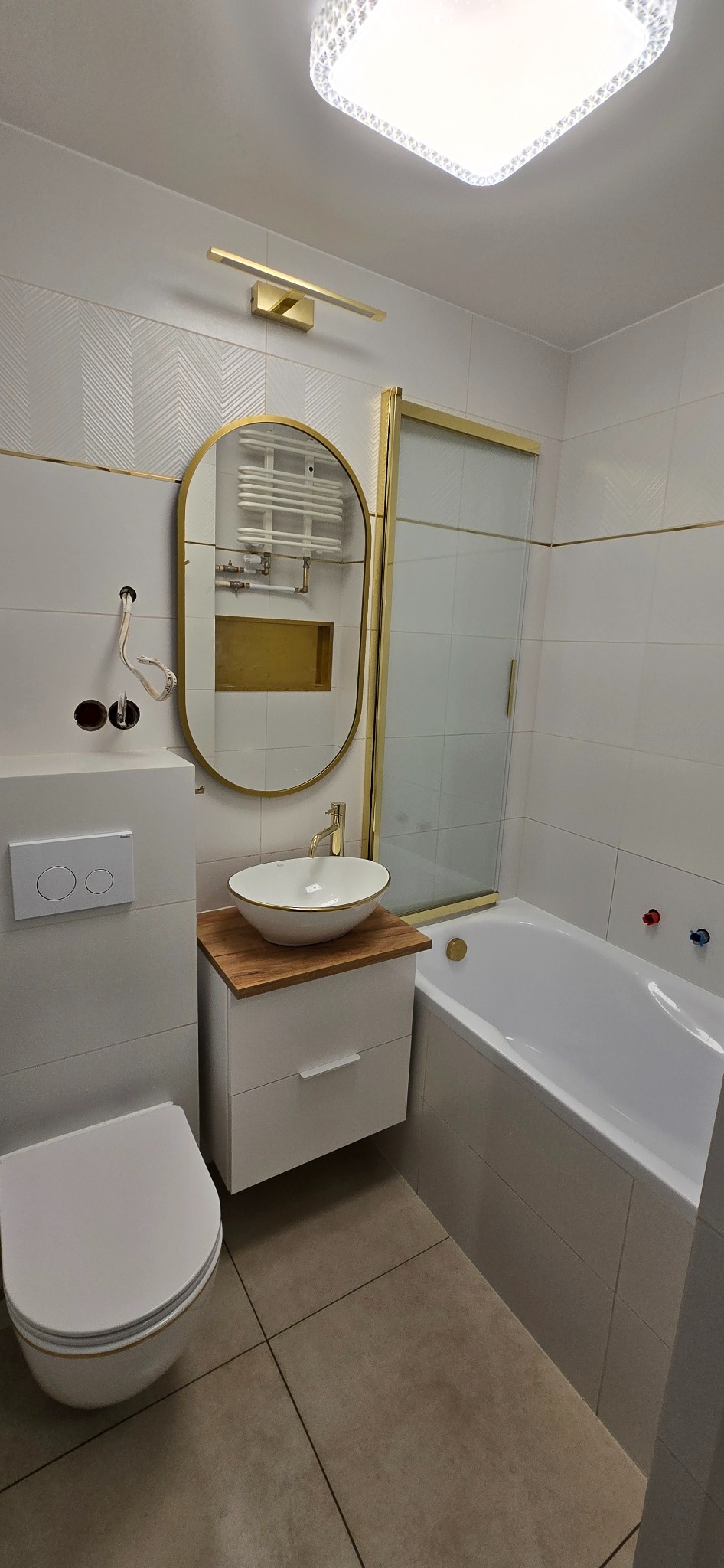 Wykończona łazienka w bieli i złocie: owalne lustro, umywalka nablatowa na szafce, kabina prysznicowa i toaleta. Widoczne niedokończone podłączenie elektryczne.