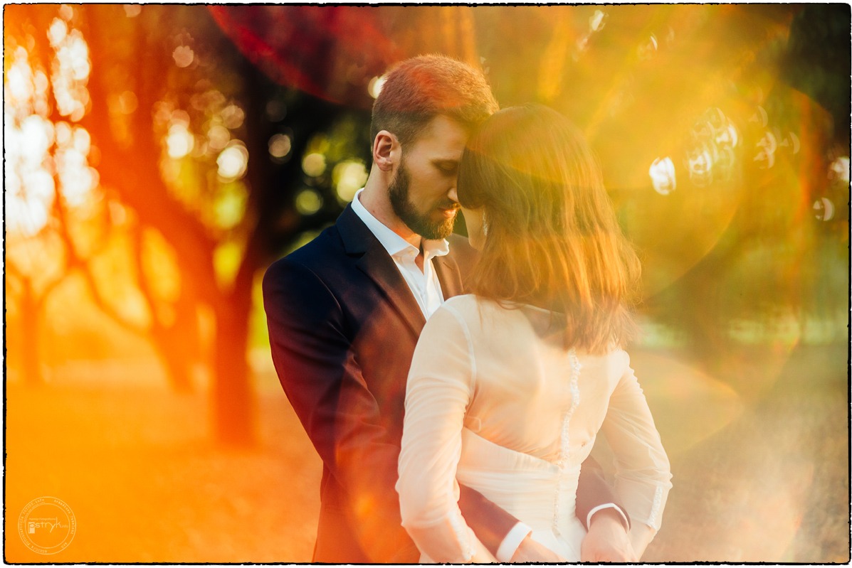 Para młoda w objęciach, on w garniturze, ona w sukni, w plenerze, z efektem bokeh i ciepłym oświetleniem. Ujęcie z bliska, skupione na emocjach i relacji.
