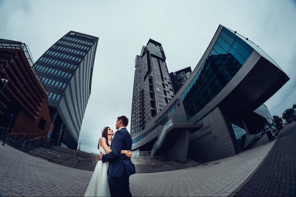 Para młoda w objęciach na tle nowoczesnej architektury miejskiej. Szeroki kąt podkreśla geometryczne kształty budynków i dodaje dynamiki kompozycji.