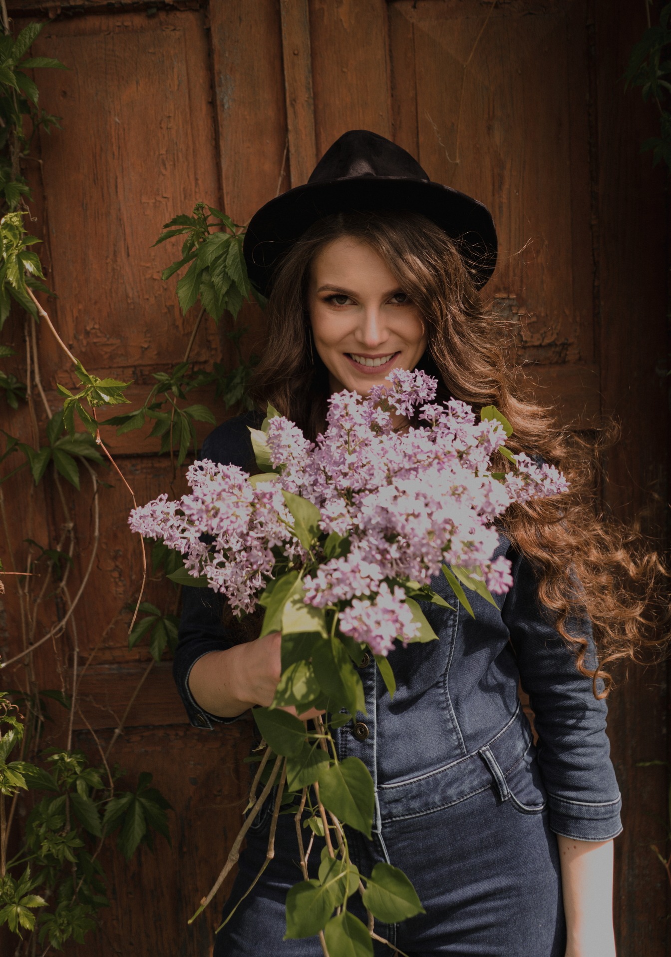 Uśmiechnięta kobieta w dżinsowym kombinezonie i kapeluszu trzyma bukiet bzów na tle drewnianych drzwi z bluszczem. Portret w naturalnym świetle.