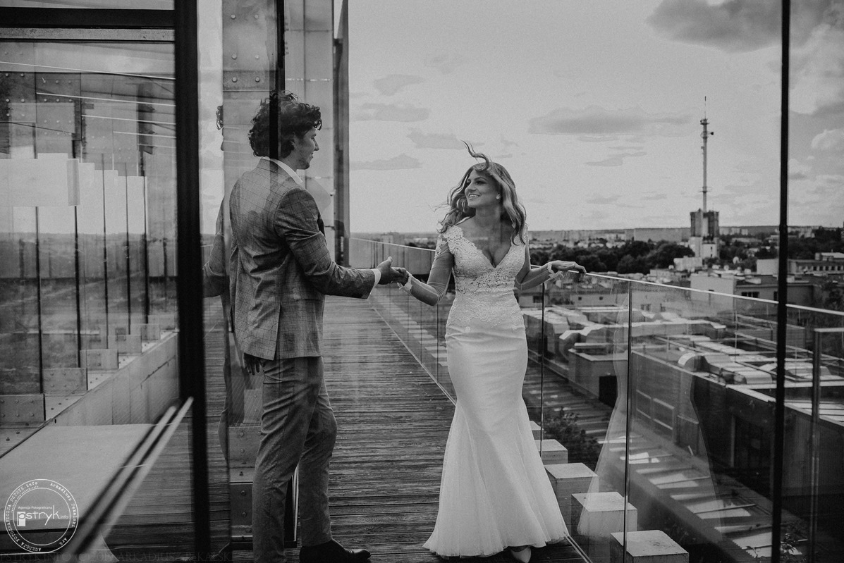 Czarno-białe zdjęcie pary w strojach ślubnych na tle miejskiej panoramy z wieżą. Ona w sukni, on w garniturze w kratę, trzymają się za ręce na tarasie z przeszkloną balustradą.