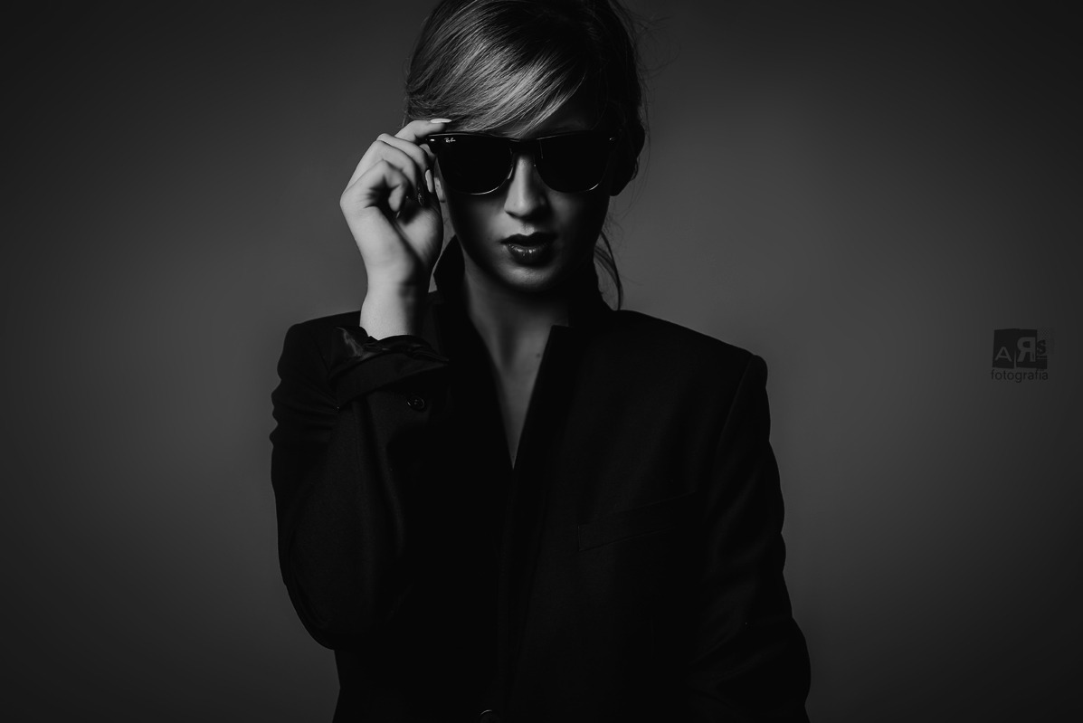 Czarno-biały portret kobiety w marynarce i okularach przeciwsłonecznych,  z delikatnym uśmiechem i wyrazistym makijażem ust, na gładkim tle.