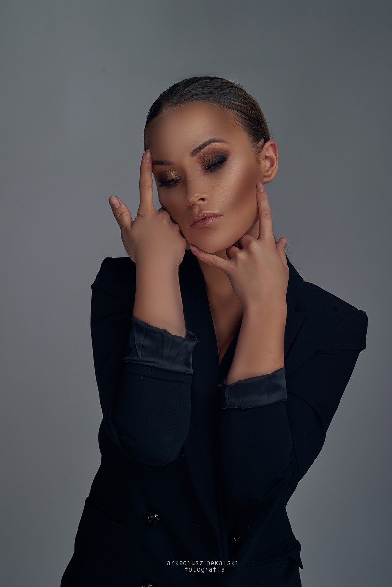 Kobieta w czarnej marynarce pozuje z dłońmi przy twarzy, makijaż w odcieniach brązu, gładko zaczesane włosy, neutralne tło, profesjonalna fotografia.