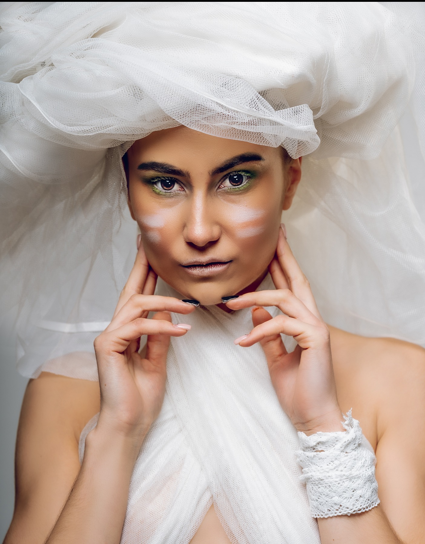 Portret kobiety z awangardowym makijażem i nakryciem głowy z tiulu, dłonie przy twarzy, artystyczna sesja w jasnej tonacji, wyraziste oczy z zielonym akcentem.