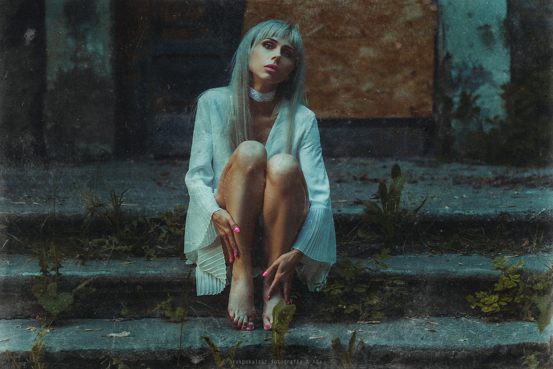 Kobieta w białej sukience siedzi na betonowych schodach, otoczona zielenią. Artystyczne ujęcie z efektem vintage i delikatnym ziarnem. Różowe paznokcie dodają koloru.