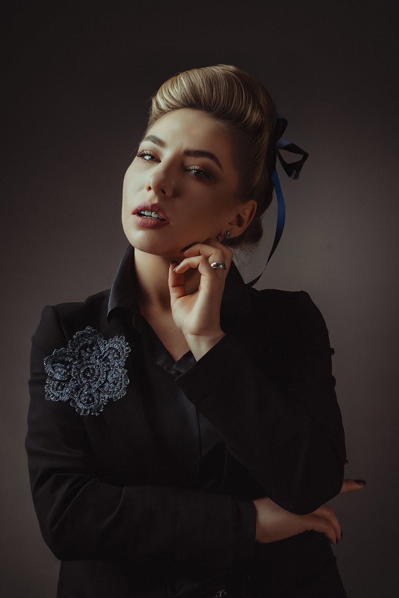 Portret kobiety w czarnej marynarce z broszką, z upiętymi włosami i niebieską wstążką, pozującej na ciemnym tle. Uwagę zwraca wyrazisty makijaż i biżuteria.