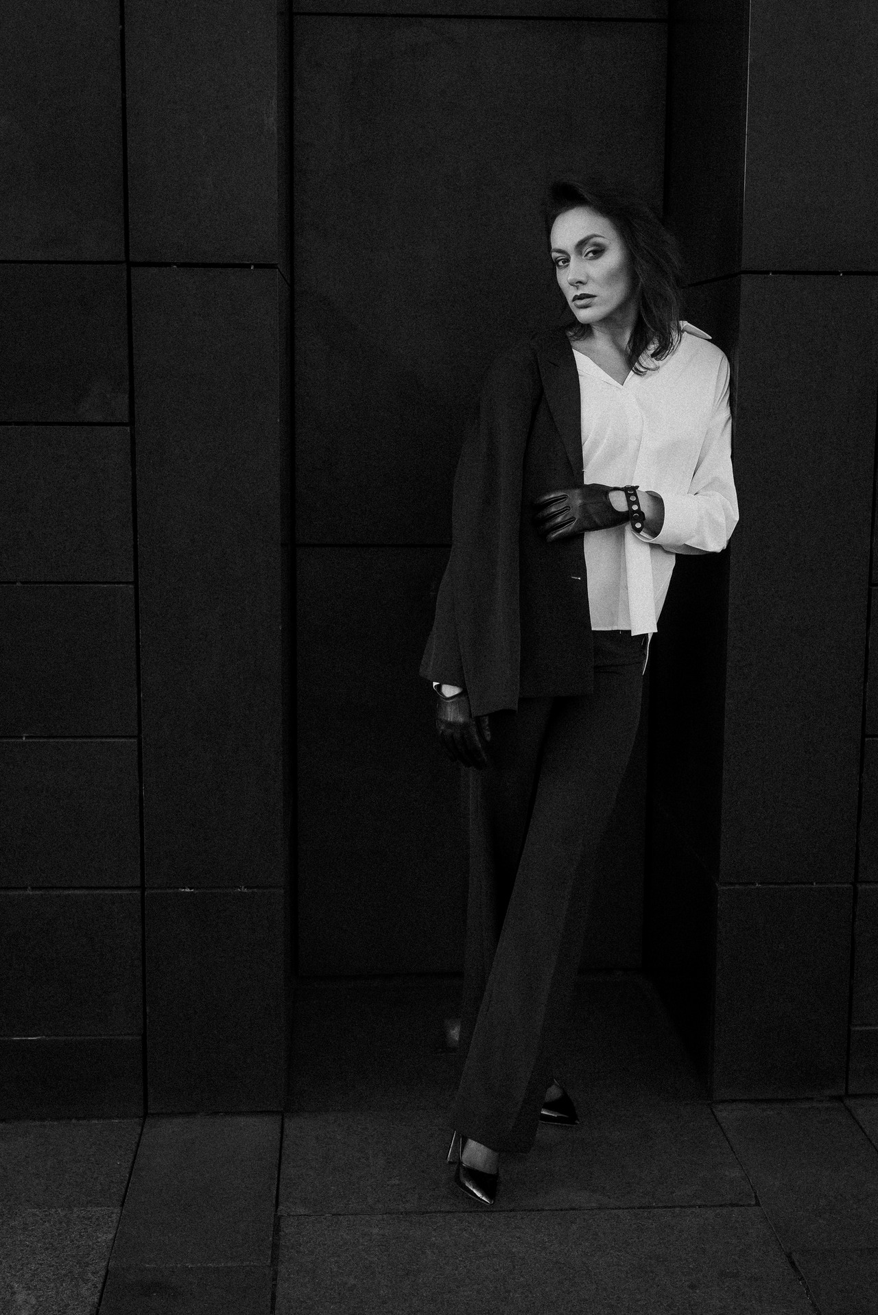 Czarno-białe zdjęcie kobiety w garniturze i białej koszuli, opierającej się o ciemną, teksturowaną ścianę. Uwagę przykuwa jej wyrazisty makijaż i skórzane rękawiczki.